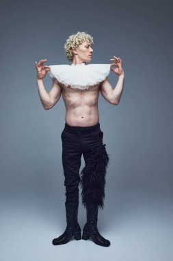 Sarışın, kıvırcık saçlı, geleneksel yakalı, sarı göğüslü genç adam, gri stüdyo geçmişine karşı dramatik bir poz veriyor. Moda birleşimi, sanat fotoğrafçılığı, kendini ifade etme. Ad