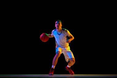 Odaklanmış atletik adam, basketbolcu tek eliyle siyah stüdyonun arka planına karşı karışık neon ışıkta top sürüyor. Profesyonel spor kavramı, enerji, güç ve güç, maç, şampiyonluk.