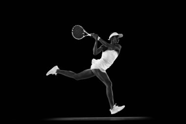 Tenis kıyafetleri içinde odaklanmış bir sporcu siyah stüdyo arka planında güçlü bir forehand vuruşu yapıyor. Monokrom filtresi. Spor anlayışı, aktif yaşam tarzları, turnuvalar ve etkinlikler, enerji, hareket.