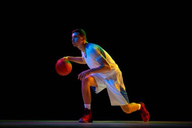 Genç, odaklanmış basketbol oyuncusu, karışık neon ışıklarıyla siyah arka planda topa karşı düşük tutuş tekniği sergiliyor. Profesyonel spor, enerji, maç, şampiyonluk kavramı.