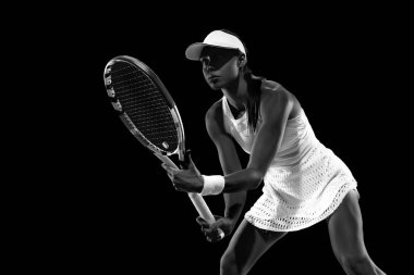 Tenis kıyafeti giymiş, rakiplerini bekleyen odaklanmış bir sporcu, siyah stüdyo geçmişine karşı hizmet veriyor. Monokrom filtresi. Kadınların spor anlayışı, aktif yaşam tarzları, turnuvalar ve etkinlikler, enerji, hareket.