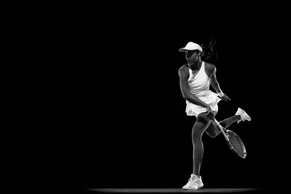 Athletic woman tennis player extending for forehand against black studio background. Monochrome filter. Concept of women in sport, active lifestyles, tournaments and events, energy, movement.