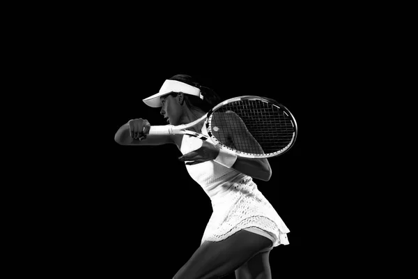 Athletic woman in tennis attire playing backhand against black studio background. Monochrome filter. Concept of women in sport, active lifestyles, tournaments and events, energy, movement.
