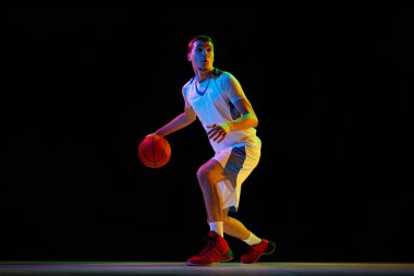 Genç, odaklanmış basketbol oyuncusu karışık neon ışıkta siyah stüdyo arka planına karşı kombine top tutma tekniği uyguluyor. Profesyonel spor kavramı, enerji, maç, turnuva, gol.