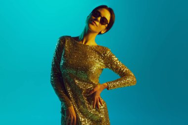 Şenlik elbisesi ve güneş gözlüğü takan güzel genç bir kadının portresi parlak sarı neon ışıkta gradyan mavi stüdyo arka planına poz veriyor. İnsan duyguları kavramı, kendini ifade etme, tarz. Ad
