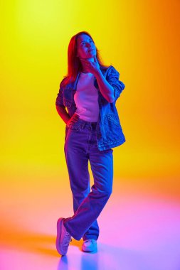 Düşünceli, rüya gören, kot giymiş genç bir bayanın portresi karışık neon ışıklarıyla değişen turuncu-sarı arka plana karşı. İnsan duyguları kavramı, kendini ifade etme, güzellik, gençlik.