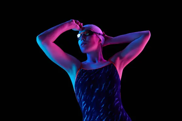 Confident, focused on competition young swimmer in swimming uniform posing in vibrant neon light against black studio background. Concept of professional sport, motion, strength and power. Ad