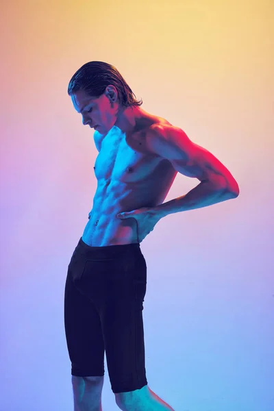 Portrait of young man with toned body posing holds hands on hips in vibrant neon light against gradient blue-pink background. Concept of natural beauty people, fitness, male health, masculinity.