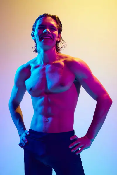 Smiling young man holds hands on hips demonstrating his perfect body curves in vibrant neon light against gradient blue-pink background. Concept of natural beauty people, male health, masculinity.