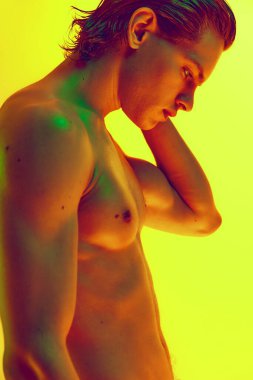 Yan görüş. Çok renkli neon ışıklı güçlü figürlü çıplak bir genç adamın, gradyan sarı arka planda çekilmiş yakın fotoğrafını çek. Doğal güzellik insanları, fitness, erkek sağlığı, erkeklik kavramı.