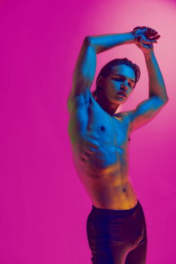 Çekici genç adam mükemmel vücut kıvrımlarını pembe stüdyo arka planına karşı karışık neon parıltısıyla nallamak için ellerini kaldırdı. Doğal güzellik insanları, fitness, erkek sağlığı ve refahı, erkeklik kavramı.