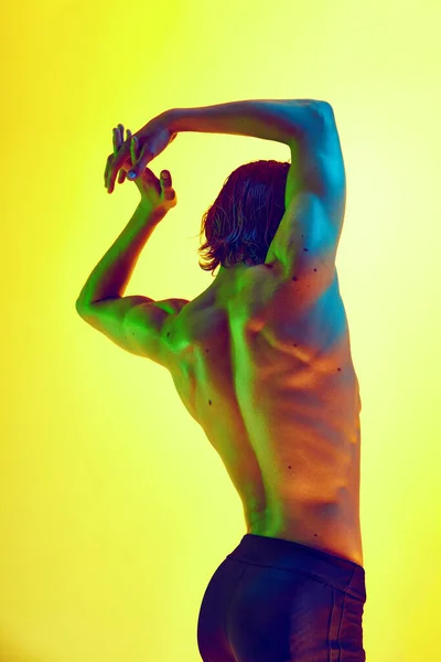 Muscular and healthy back. Man posing in multicolored neon light against gradient yellow background. Concept of natural beauty people, fitness, male health and wellness, masculinity.