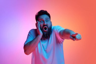 Vay canına. Şok olmuş genç bir adam, ağzı açık bir şekilde elini yüzüne koydu ve neon ışıkta, canlı gradyan stüdyo arkaplanına karşı mesafeyi gösterdi. İnsan duyguları kavramı, kendini ifade etme, moda.