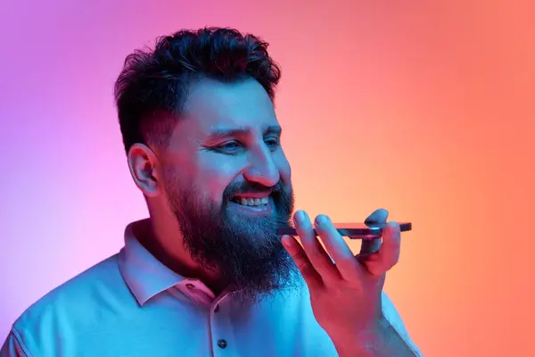 Man holding phone to mouth while talking on speaker phone in neon light against vibrant gradient studio background. Concept of human emotions, self-expression, connection in distance, delivery. Ad