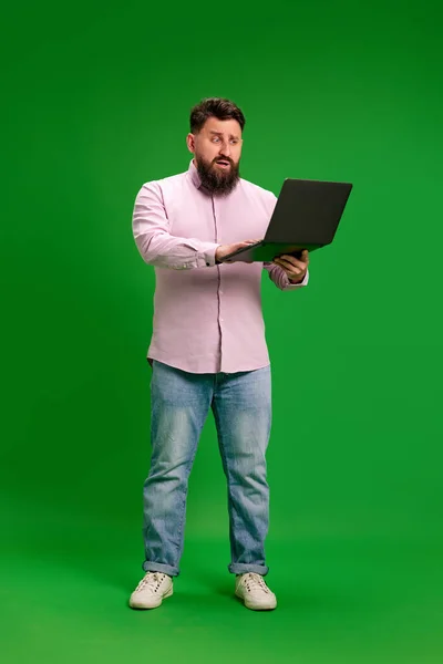 Full-length portrait of young man with beard looks at laptop screen with blank look against vibrant green studio background. Concept of human emotions, self-expression, work, study, network connection