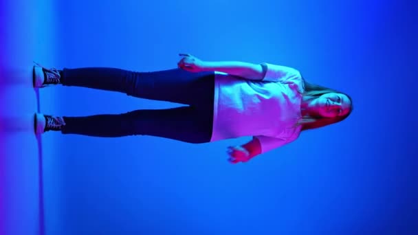 穿着休闲装的快乐而积极的女人在霓虹灯下与渐变的蓝色工作室背景跳舞 美的概念 自我表达 人类的情感 垂直方向 — 图库视频影像