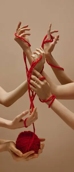 Unity. Womans hand holds ball of red threads and other hands intertwined with each other tied with this thread against sandy color studio background. Concept of human touch, beauty and care.