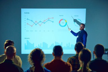 Orta yaşlı genç adam, analitik bilgilerle konferans odasında öğrencilerin konuşmasını dinlediği ekranın yanında duruyor. İş kavramı, girişim, liderlik kişisel gelişim kursları.