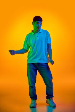 Sıradan giyimli Asyalı genç bir adam, kolları açık bir dansçı, parlak neon ışıklarıyla rengarenk turuncu arka planda yıkanmış. İnsan duyguları, moda ve güzellik kavramı, kendini ifade etme, çalışma, hobi.