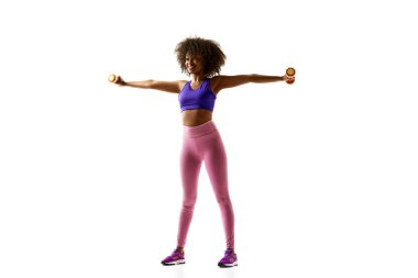 Mavi ağırlıklarla egzersiz yapan neşeli Afro-Amerikalı kadın, beyaz stüdyo arka planına karşı mor spor kıyafetleri giyiyor. Spor anlayışı, yas tutma alışkanlığı, aktif ve sağlıklı yaşam tarzı, enerji..