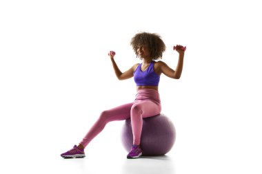 Mor spor giysili Afro-Amerikalı bir kadın egzersiz topunun üzerinde oturuyor, beyaz stüdyo arka planına karşı küçük ağırlıklar kaldırıyor. Spor anlayışı, yas tutma alışkanlığı, aktif ve sağlıklı yaşam tarzı, eylem.