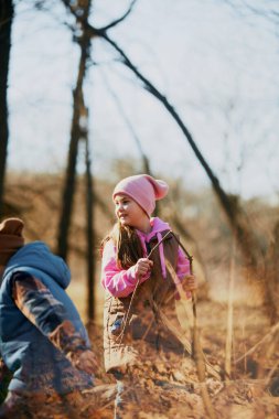 Sıcak ceketli ve şapkalı küçük çocuklar ağaçlarla çevrili basit bir sopayla neşe buluyorlar. Çocuk gelişimi, okul, çocukluk, moda ve tarz için açık hava aktiviteleri kavramı. Ad