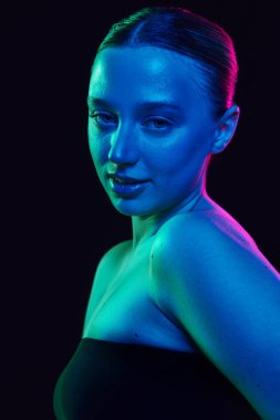 Çıplak omuzlu, siyah omuzlu genç bir kadın siyah stüdyo arka planına karşı neon ışıkta kameraya bakıyor. Sağlık, spa prosedürleri, kozmetoloji, yaşlanma ve cilt bakımı ürünleri kavramı.