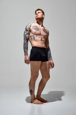 Spor vücudunda dövmeleri olan, gri stüdyo geçmişine karşı iç çamaşırlarıyla poz veren genç, çekici, üstsüz bir adam. Doğal güzellik, moda ve tarz kavramı, erkek sağlığı ve vücut bakımı. Ad