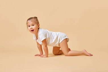 Küçük güzel bir bebeğin yan görüntüsü, yerde sürünen bir kız tatlı beyaz bir tulum içinde pastel renkli arka planda. Çocukluk kavramı, annelik, yaşam, doğum. İlan için alanı kopyala