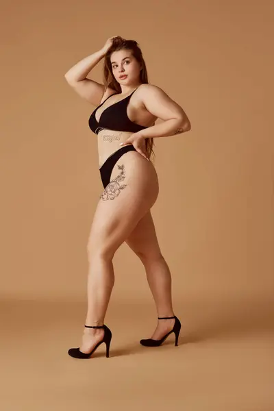 Full length portrait of young chubby woman posing in lingerie standing on heels against sandy color studio background. Self-expression. Concept of natural beauty, femininity, body positivity, spa.