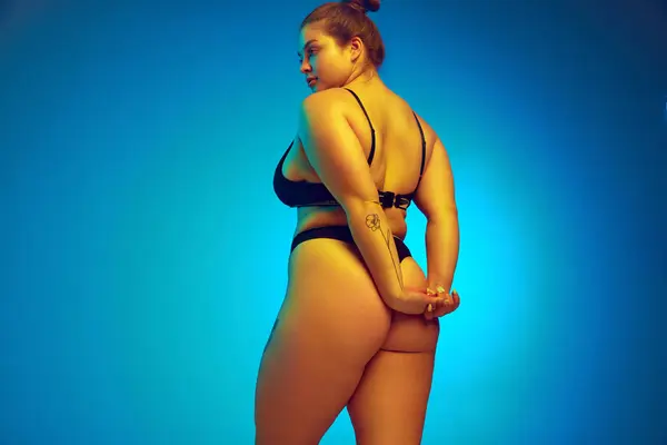 Rear view portrait of plus-size female model posing holds hands behind back in yellow neon light against blue studio background. Concept of natural beauty, femininity, body positivity, diet, spa.