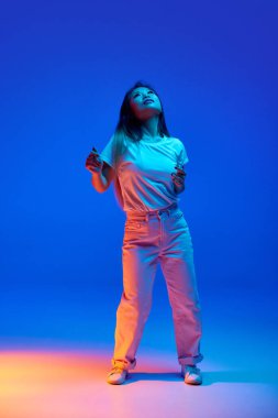 Neşeli, genç Asyalı kadın resminin tam boy portresi gündelik kıyafetlerle dans ediyor, mavi arka planda neon ışıkta el kaldırıyor. İnsan duyguları, kendini ifade etme, dans ve müzik kavramı.