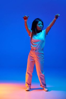 Neşeli, genç Asyalı kadın resminin tam boy portresi gündelik kıyafetlerle dans ediyor, mavi arka planda neon ışıkta el kaldırıyor. İnsan duyguları, kendini ifade etme, dans ve müzik kavramı. Ad