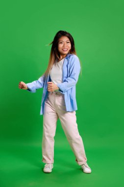 Genç ve çekici Asyalı bir kadının canlı yeşil stüdyo arka planında günlük kıyafetlerle dans edişinin tam boy portresi. İnsan duyguları, kendini ifade etme, rahatlama, eğlence ve neşe kavramı. Ad