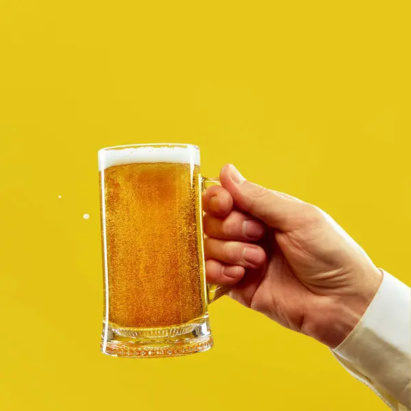 男性手拿着冰凉的杯子 泡沫般的啤酒 金色的啤酒 背景鲜明的黄色工作室 Oktoberfest 度假的概念 广告的复制空间 免版税图库图片