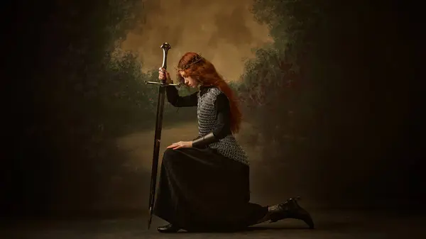 多愁善感的战士公主 女人跪在剑下 面对着古老的背景 摆出沉思的姿势 历史与现代的概念 美与时尚 免版税图库照片