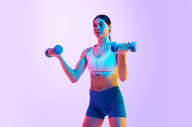 Genç atlet kadın, dambıllar için neon ışıklarıyla egzersiz yapıyor. Gradyan stüdyo arka planına karşı. Spor ve eğlence anlayışı, hareket, kişisel bakım, eylem, enerji. Ad