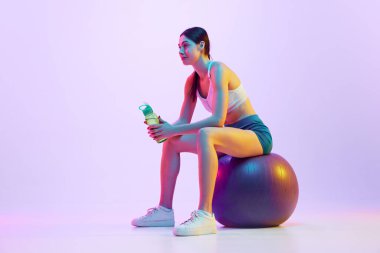 Eğitimler arasında dinlenen genç bir kadın. Stüdyonun arka planına karşı neon ışıkta su savaşıyla uyumlu bir topun üzerinde oturuyor. Spor ve eğlence kavramı, hareket, kişisel bakım, eylem, enerji.