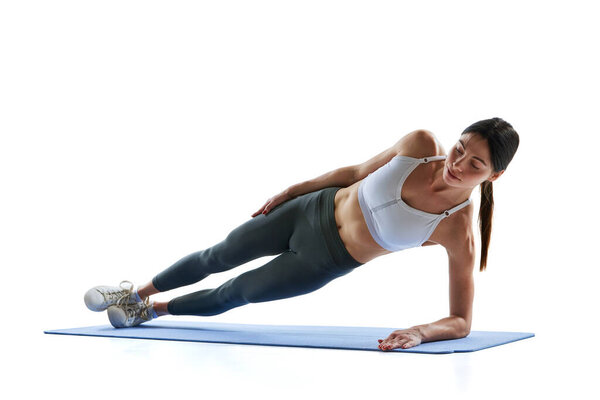 Dynamic portrait of young athlete woman stands in side plank against white studio background. Abdominal muscles. Concept of sport and recreation, movement, self care, action, energy. Ad