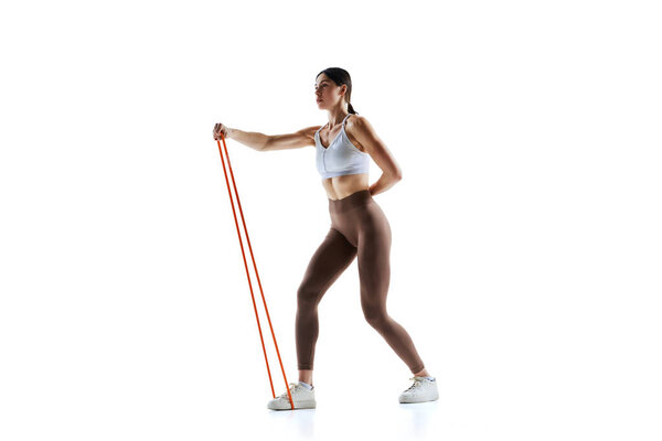 Fit female athlete in fashion top and leggings demonstrating use of exercise band against white studio background. Concept of sport and recreation, movement, self care, action, energy. Ad