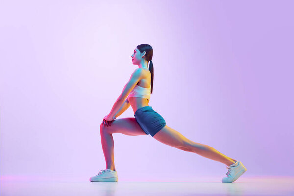 Young athlete woman doing lunges in motion in neon light against gradient studio background. Side view portrait. Concept of sport and recreation, movement, self care, action, energy. Ad