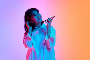 Düşünceli genç bir kadının portresi. Neon ışıkta telefonla konuşurken saçlarına bakıp dokunuyor. Gradyan stüdyo arka planına karşı. İnsan duyguları kavramı, moda, güzellik, tarz.