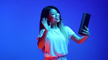 Gülümseyen Asyalı kadın dijital tablette neon ışıkta mavi gradyan zemin karşısında video toplantısı düzenlerken el sallıyor. İş, girişim, satış, çalışma ve öğrenim kavramı.