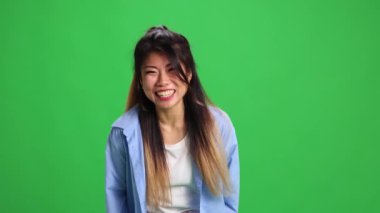 Genç, güzel Asyalı bir kadın, günlük kıyafetli bir öğrenci canlı yeşil stüdyo arka planında kameraya bakıp gülüyor. İnsan duyguları kavramı, kendini ifade etme, moda, tarz.