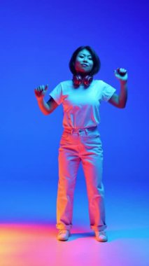 Neşeli genç Asyalı kadın, günlük kıyafetleriyle dans ediyor, neon ışıkta mavi renk değişimine karşı ellerini kaldırıyor. İnsan duyguları, kendini ifade etme, gençlik ve güzellik, dans ve müzik kavramı.