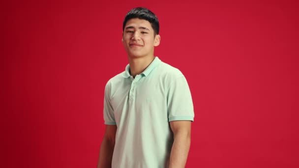 若くてハンサムなアジアの男性は 赤いスタジオの背景に対してカメラを見ながら笑顔でカジュアルな服を着ています 人間の感情 ファッション 美しさ 自己表現 趣味の概念 アドベンチャー — ストック動画