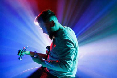 Neon ışıkta elektro gitar çalan genç bir adamın yan görüş portresi ve sahnede görünür bir duman. Rock ve klasik müzik kavramı, hobi ve iş, enerji, müzik festivalleri, konserler. Ad