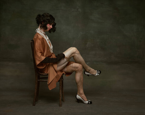 Side view portrait of young man in dress sitting on wooden chair and fitting stockings against vintage studio background. Concept of self-expression, comparisons of eras, freedom, human rights.