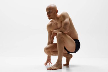 Çömelmiş iç çamaşırlarıyla poz veren zinde genç bir adam, gri stüdyo geçmişine karşı odak ve atletik formunu gösteriyor. Erkek sağlığı, kişisel bakım, moda ve güzellik kavramı, sağlıklı yaşam tarzı. Ad