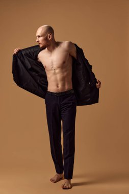 Gömleksiz genç adam zarif bir pantolon ve ceket giydi. Stüdyonun arka planına karşı gelişmişlik gösteriyordu. Erkek sağlığı, kişisel bakım, moda ve güzellik kavramı, sağlıklı yaşam tarzı